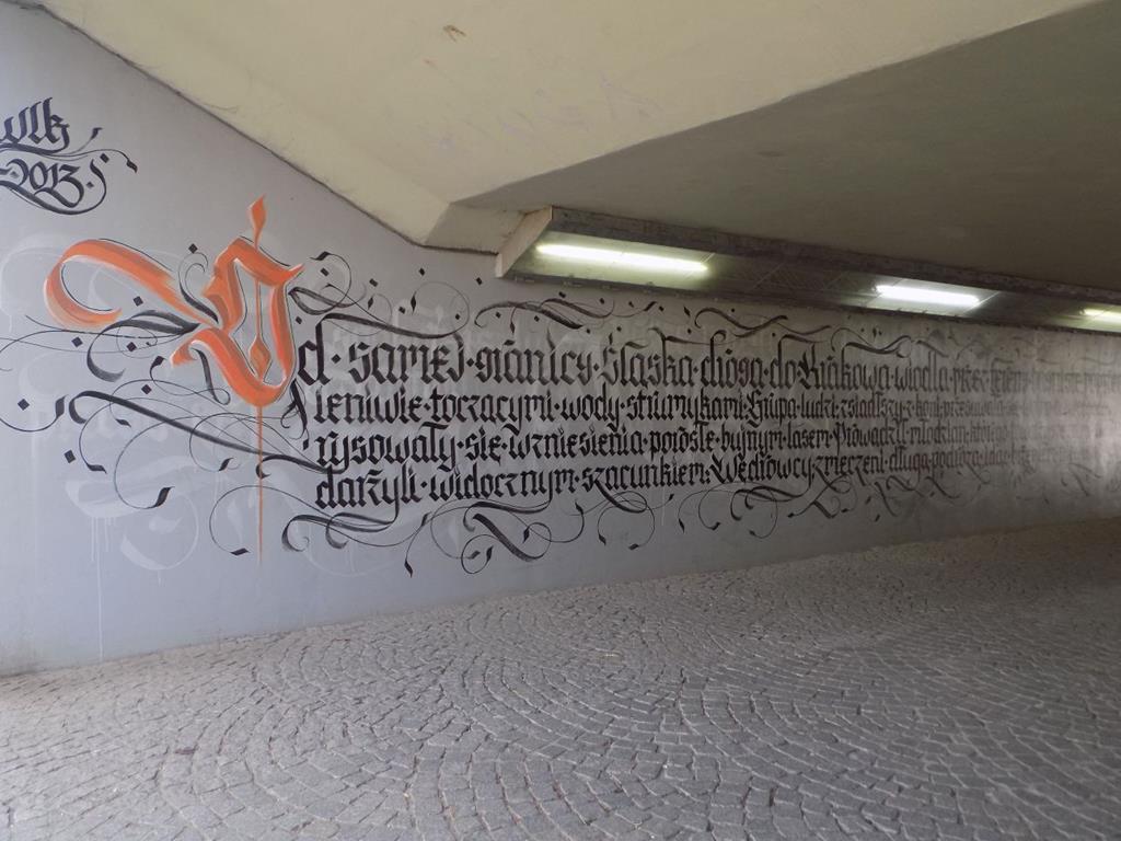 Mural na szarym tle w tunelu, czarne napisy, pierwsza litera duża oraz pomarańczowa