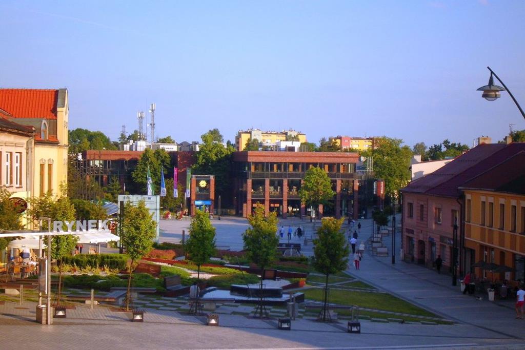 Różno-kolorowe budynki, po lewej stronie duży biały napis: "rynek", koło niego oraz wokół budynków drzewa