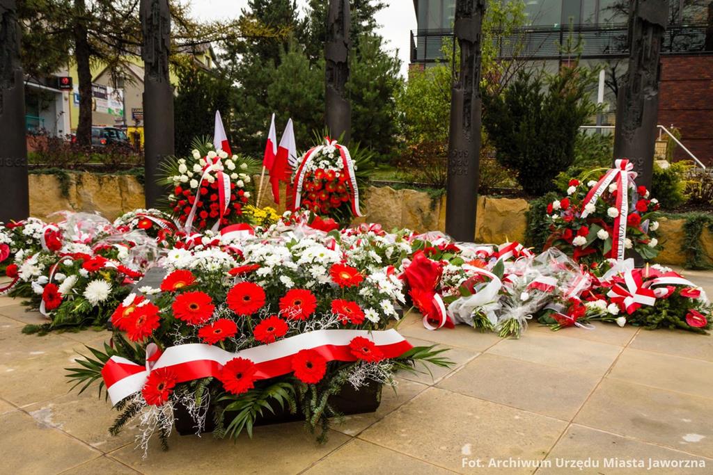 Biało-czerwone kwiaty, z wstążkami flagi Polski oraz flagi Polski przy pomniku