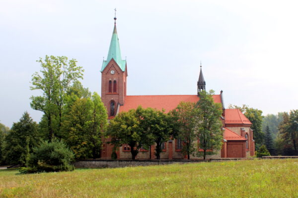 Szaro-czerwony kościół z dwoma wieżami: jedna mniejsza czarna, druga miętowa, obydwie są zakończone czarnym krzyżami, obok kościoła stoją drzewa