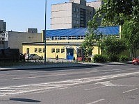 Fragment ulicy i chodnika przy żółtym budynku hali sportowej, w oddali bloki i drzewa. 