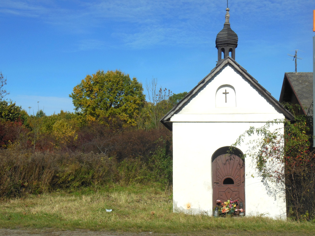 Niebieskie niebo, zielone zarośla, biała kapliczka z brązowymi drzwiami