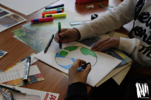Uczestnicy warsztatów podczas pracy: zbliżenie na dłonie, rysowanie flamastrami, wokół rozłożone na stole gazety i materiały plastyczne.