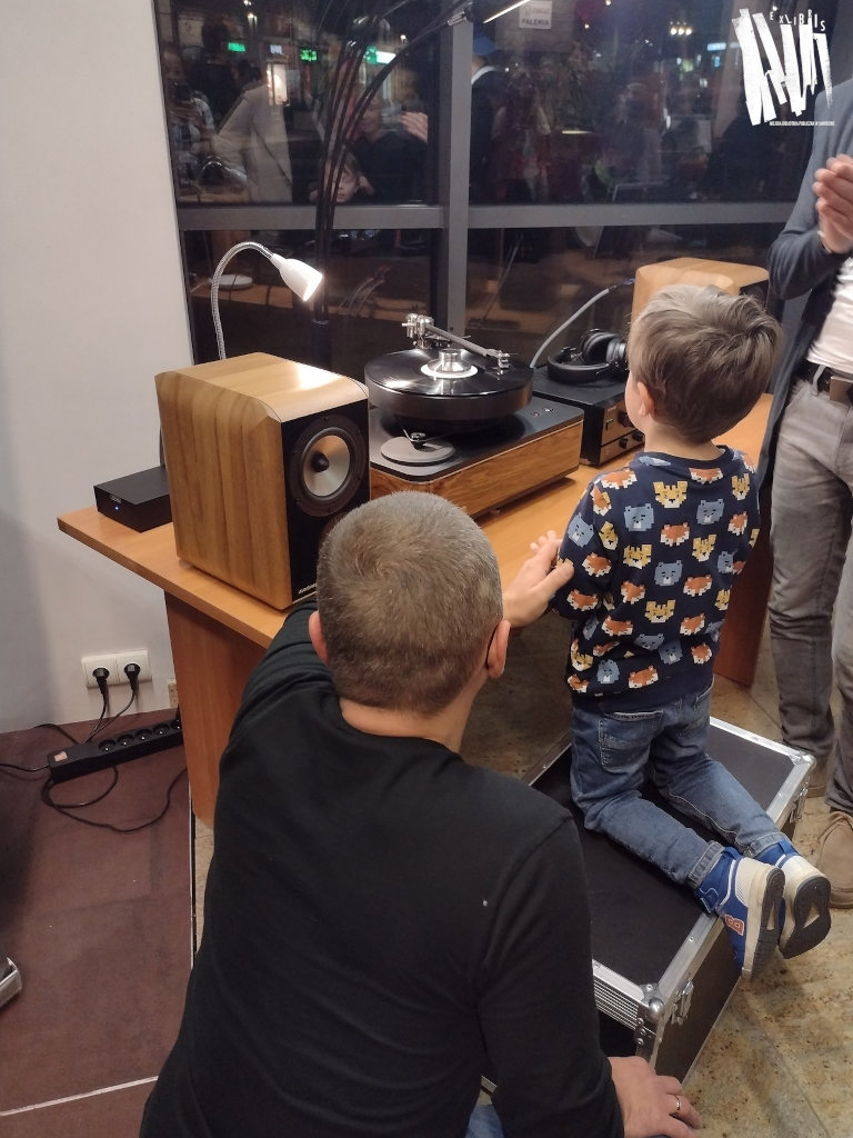 Na stoliku koloru pomarańczowego stoi gramofon i kolumny. Przed stolikiem, na skrzyni klęczy mały chłopieć wpatrujący się w sprzęt. Tuz obok niego, tyłem do robiącego zdjęcie przykucnął ojciec malucha.