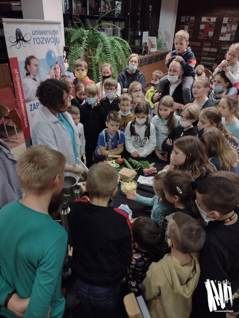 Zdjęcie przedstawia grupkę dzieci, otaczających stolik, na którym umieszczone są materiały do przeprowadzenia eksperymentów chemicznych.