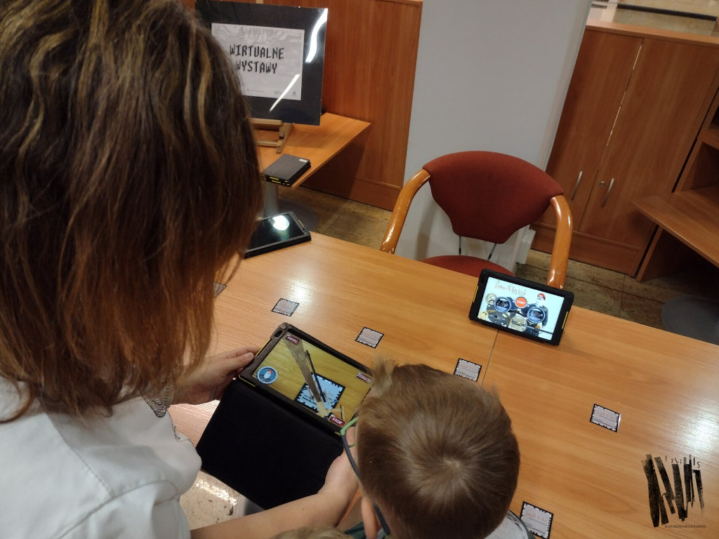 Dwa stoliki koloru pomarańczowego, na których poukładane są kody QR i tablety. Na pierwszym planie widać głowy dwóch osób wpatrzonych w Ekron tabletu.