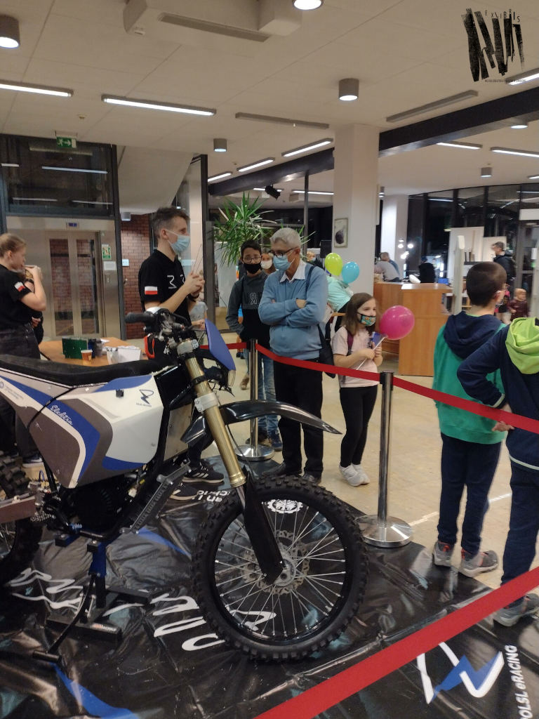 Wnętrze pomieszczenia, białe, wysokie sufity. Na pierwszym planie stoi motor, odgrodzony od grupy oglądających czerwoną taśmą.