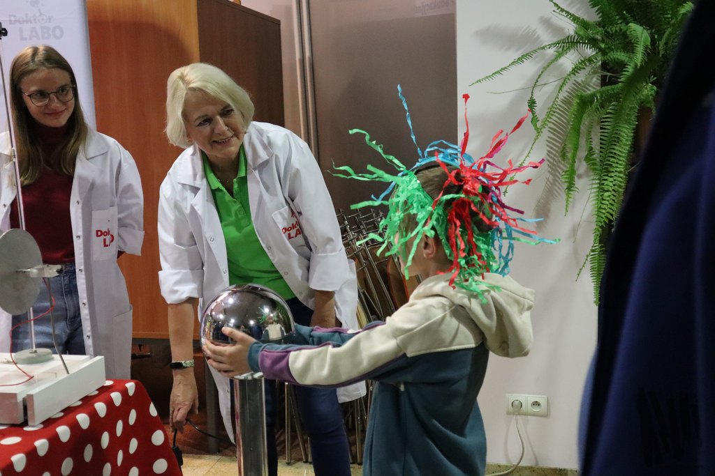 Niedaleko stołu nakrytego czerwonym obrusem stoi dziecko ubrane w kremowo-granatowo-fioletową bluzę. Uczestnik został uwieczniony w trakcie doświadczenia wykonywanego z pomocą ekspertów firmy Doktor Labo. Chłopiec trzyma dłonie na dużej kuli stojącej przed nim, a na głowę ma założony chełm z przyczepionymi do niego paskami kolorowej bibuły, które sterczą na wszystkie strony. Tuż obok uczestnika stoją dwie kobiety w białych fartuchach – długowłosa szatynka w okularach oraz starsza blondynka – które czuwają nad przebiegiem doświadczenia i bezpieczeństwem uczestnika.
