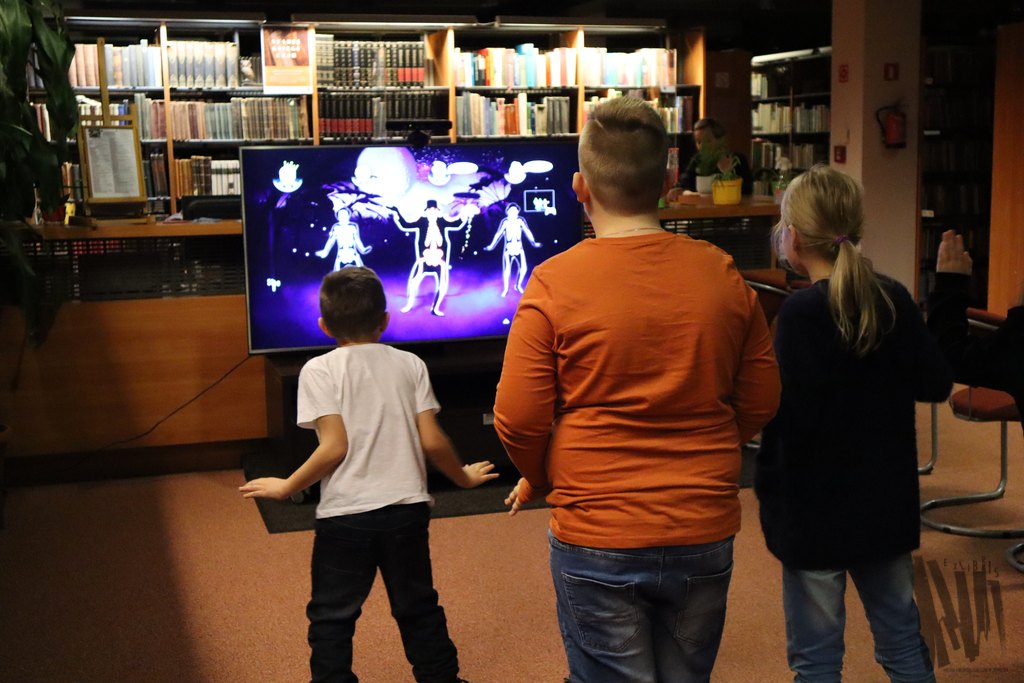 W Czytelni Naukowej, w centrum fotografii, widać duży ekran, na którym wyświetlona jest gra na konsolę XBOX 360 przedstawiająca tańczące szkielety. Przed ekranem znajduje się troje młodych uczestników uchwyconych w trakcie ruchu, gdyż nie grają za pomocą padów, ale ruchów własnego ciała. Pierwszy chłopiec, który pochyla się w stronę ekranu, jest najdrobniejszy, ma krótkie brązowe włosy, a ubrany jest w białą koszulkę i czarne spodnie. Tuż obok stoi drugi, dużo wyższy chłopiec o krótkich blond włosach, ubrany w pomarańczową bluzkę i niebieskie dżinsy. Na końcu stoi dziewczynka o bardzo jasnych włosach spiętych w kucyk, która ubrana jest w czarną bluzeczkę i niebieskie dżinsy.