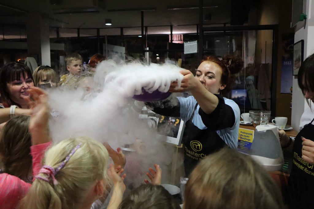 Przy stanowisku Uniwersytetu Rozwoju z Chorzowa dwie kobiety w czarnych fartuchach ze złotym logo Masterchef prowadzą pokaz kuchni molekularnej. Stanowisko otacza tłum młodych uczestników, na twarzach których maluje się podziw i zachwyt, a ich dłonie wyciągają się w stronę dymu z ciekłego azotu. Ów dym unosi się z fioletowej misy, którą wysoko nad głowę podnosi rudowłosa kobieta w czarnym fartuchu, znajdująca się w centrum fotografii.
