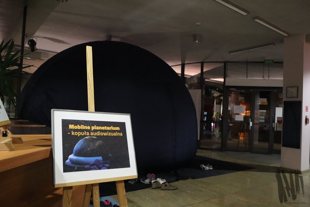 Na przestrzeni między wejściem do Biblioteki a Galerią ExLibris widać namiot mieszczący mobilne planetarium służące do pokazów dla dzieci. Planetarium rozłożono na czarnej podkładce, a jego kształt przypomina czarną półkulę. Wokół planetarium, na czarnej podkładce ustawiono dziecięce buty, a nieco bliżej kamery znajduje się tabliczka z wizerunkiem planet i napisem „mobilne planetarium – kopuła audiowizualna”.