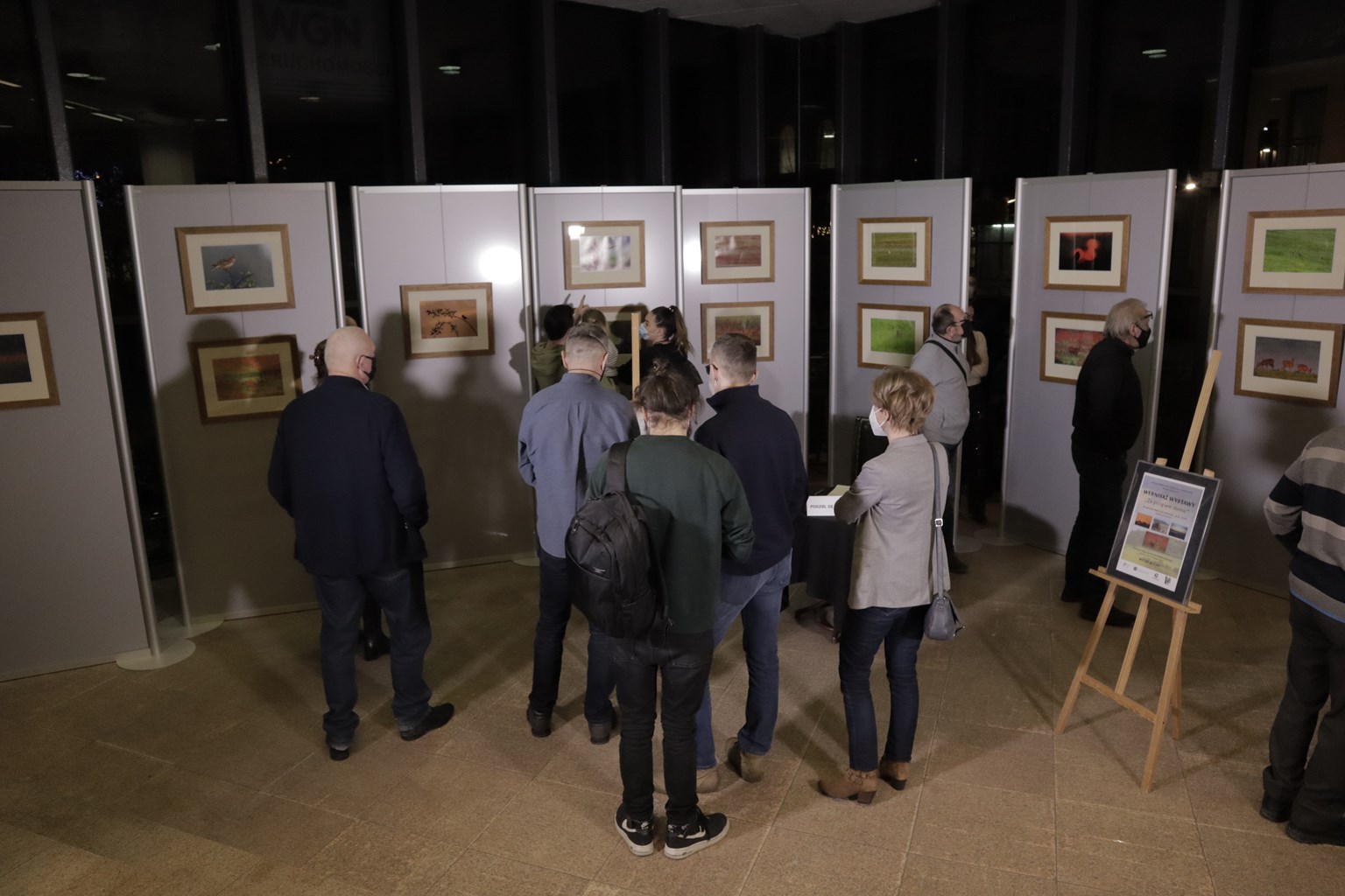 Uczestnicy wernisażu: siedmiu mężczyzn, trzy kobiety i kilkuletnia dziewczynka, stoją zwróceni tyłem do kamery, podziwiając fotografie prezentowane w galerii ExLibris.