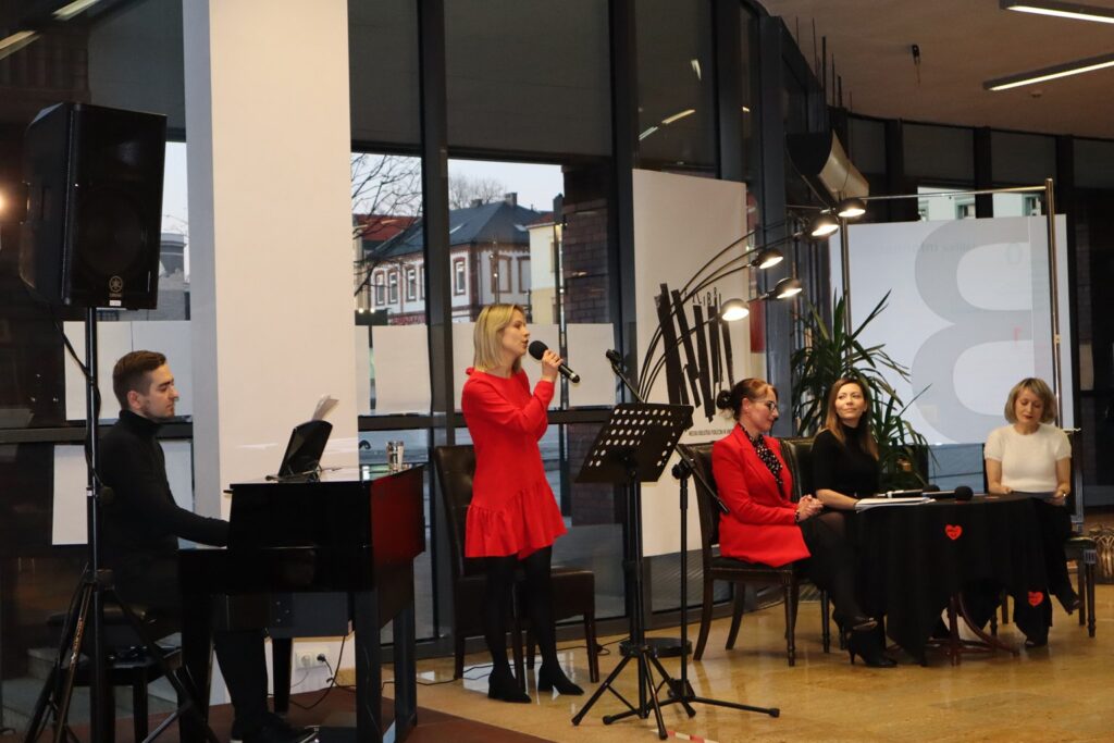 Hol Biblioteki Głównej, na scenie, po lewej stronie przy pianinie siedzi muzyk, Piotr Laska, obok stoi wokalistka Marzena Rendak i śpiewa do trzymanego w ręce mikrofonu. Po prawej stronie siedzą przy okrągłym stoliku trzy kobiety.