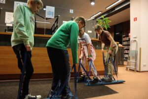Na terenie Wypożyczalni dla Dorosłych mijają się dwie pary uczestników, próbujących się poruszać na dwuosobowych nartach, sterowanych za pomocą trzymanych w dłoniach szelek. Pierwsza para to chłopcy, a druga dziewczęta – oba zespoły wykonują zadanie z malującą się na twarzach mieszanką skupienia i rozbawienia.
