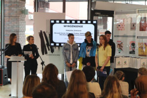 Zbliżenie na scenę, na której wciąż stoi zespół laureatów z Ukrainy. Chłopcy trzymają w dłoniach torby z nagrodami, a towarzysząca im kobieta przemawia do mikrofonu. Na twarzach wszystkich obecnych na scenie: laureatów, opiekunów i prowadzących widać powagę i smutek związany z sytuacją panującą na Ukrainie.
