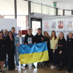 Zdjęcie grupowe, w centrum którego znajduje się wyróżniony zespół z Ukrainy. Trzech nastolatków trzyma przed sobą rozłożoną flagę Ukrainy, a z obu stron otaczają ich pracownicy biblioteki, w znakomitej większości ubrani na czarno.