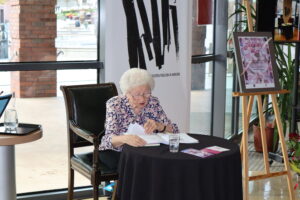 Na kolorowym zdjęciu widzimy poetkę Alicję Dudek siedzącą przy okrągłym stoliku przykrytym czarnym obrusem. Za autorką widzimy dużo logo biblioteki.
