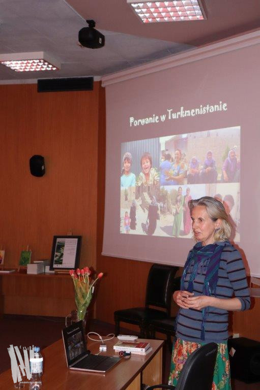 Audytorium Biblioteki Głównej w Jaworznie przy biurku stoi prowadząca Marzena Filipczak. Obok duży ekran, na którym widnieje kadr z prezentacji multimedialnej, przygotowanej przez autorkę z widocznym tytułem spotkania Porwanie w Turkmenistanie.
