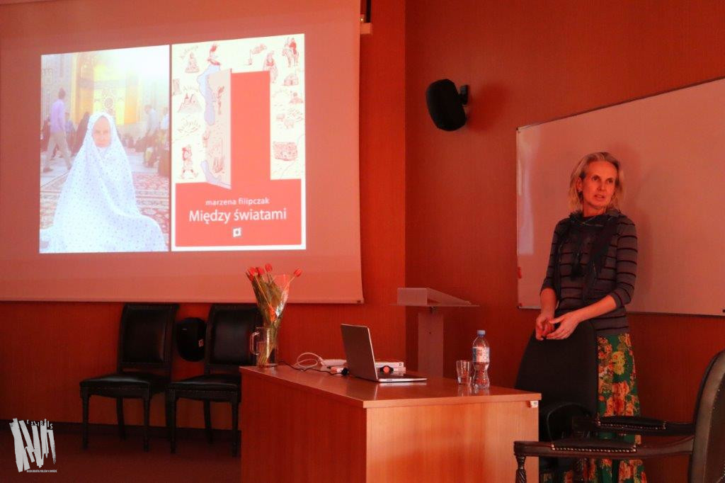 Audytorium Biblioteki Głównej w Jaworznie przy biurku stoi prowadząca spotkanie Marzena Filipczak. Obok duży ekran, na którym widnieje kadr z prezentacji multimedialnej, przygotowanej przez autorkę.