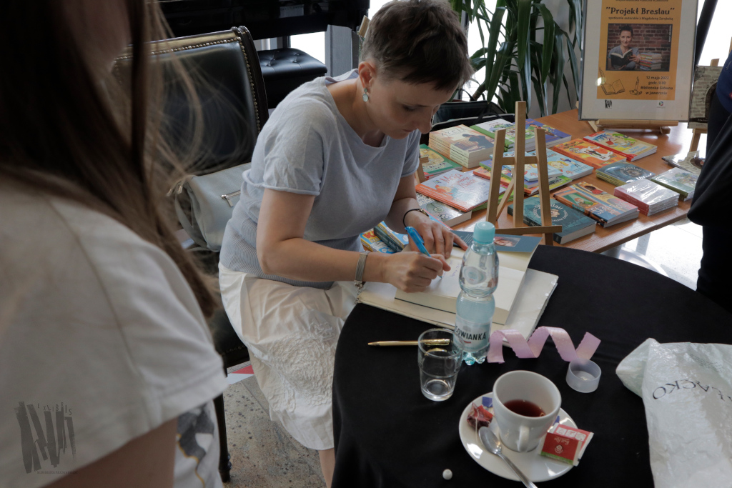 Przy okrągłym stoliku, nakrytym czarnym obrusem, siedzi Magdalena Zarębska. Autorka pochyla się nad otwartą książką, w której pisze dedykację i składa autograf dla, stojącej tyłem do kamery, dziewczynki o długich brązowych włosach.