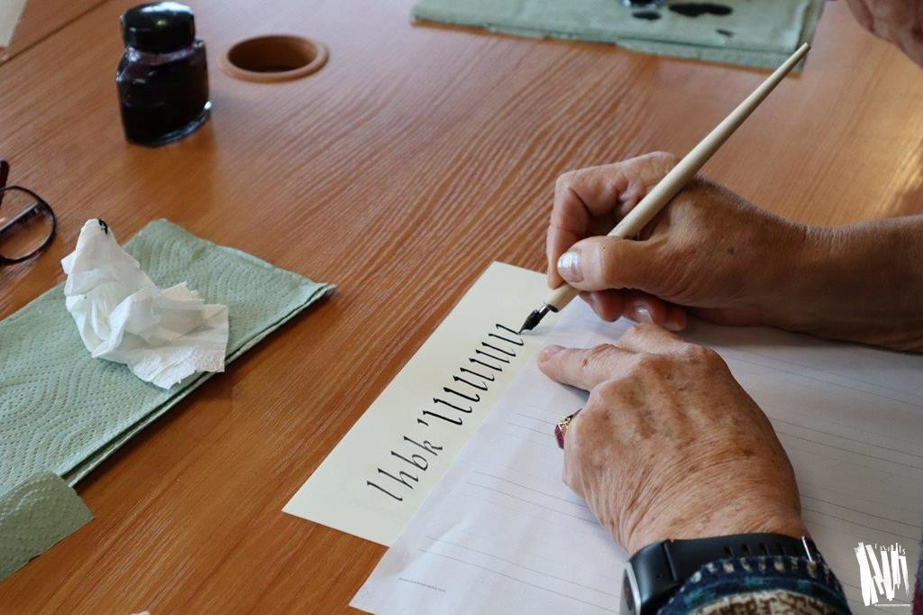 Zbliżenie na dłonie jednej z seniorek, która kończy pisać pierwszą linijkę liter na kartce A4. Kobieta pisze piórem i czarnym tuszem, którego słoiczek stoi nieopodal.