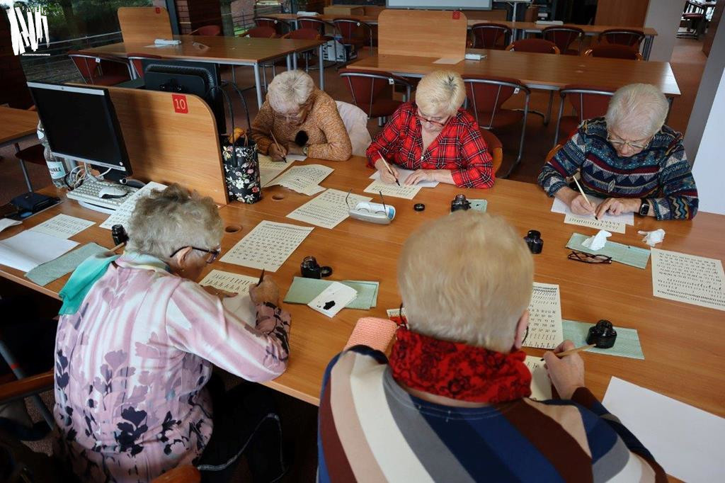 Przy stole w Czytelni Naukowej siedzi sześć siwowłosych kobiet. Stół jest zastawiony słoiczkami z atramentem oraz materiałami do ćwiczeń. Wszystkie seniorki pochylają się nad kartkami papieru, które zapełniają wykaligrafowanymi znakami, tworzonymi przy pomocy specjalnych piór i atramentów.