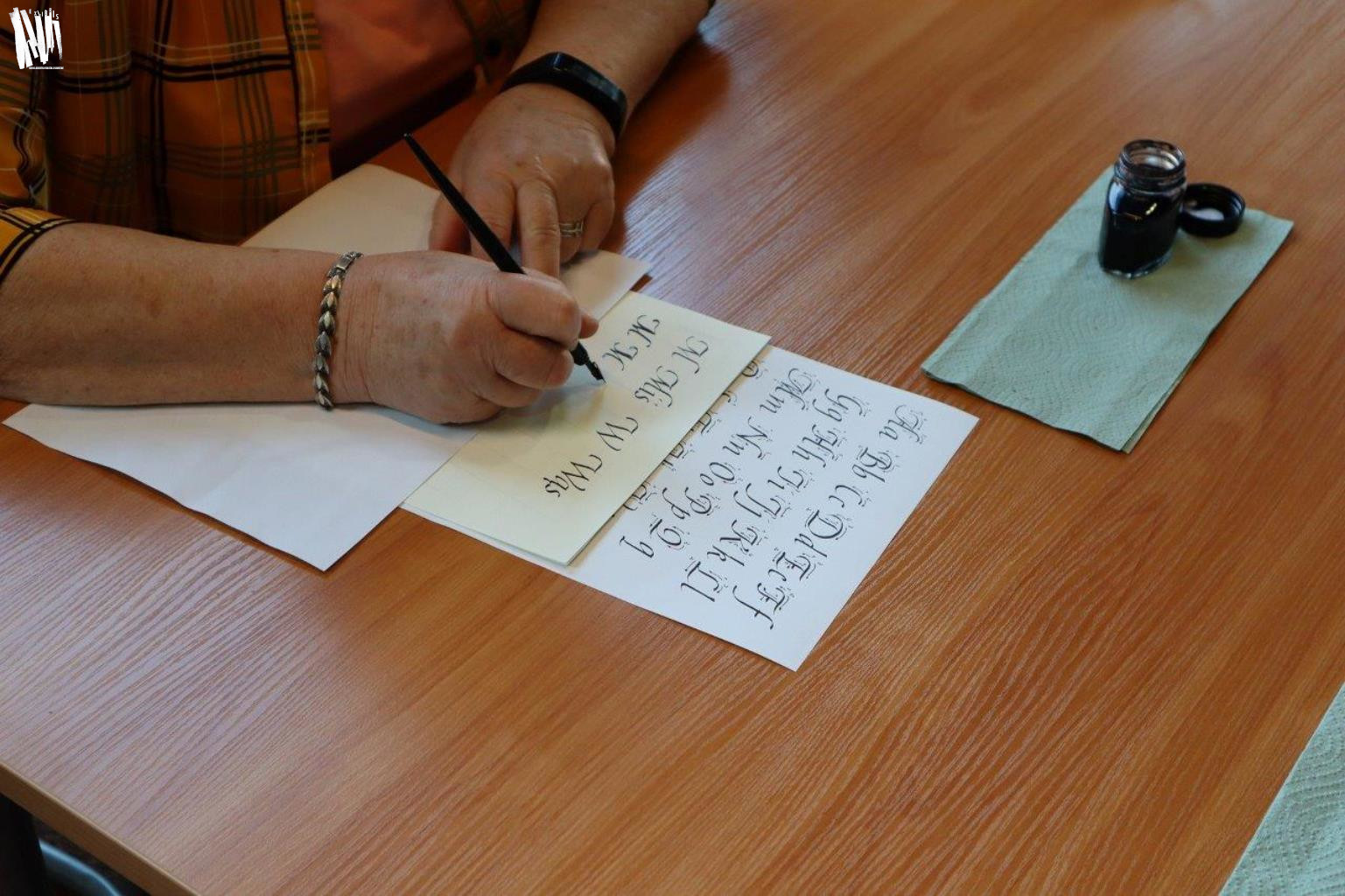 Osoba siedząca przy stole ćwiczy pisownię liter według wzornika. Zbliżenie na dłonie. W jednej z nich uczestnik warsztatów trzyma pióro. Obok na papierowej serwetce stoi otwarty kałamarz.