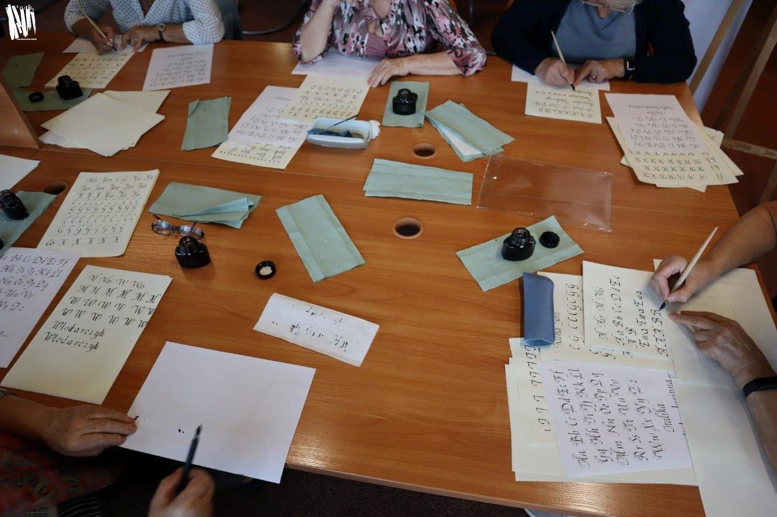 Stół w Czytelni Naukowej zastawiony materiałami do kaligrafii - kałamarzami z atramentem i kartkami papieru do ćwiczeń. Przy stole siedzi pięć osób, każda z nich trzyma w ręce pióro.