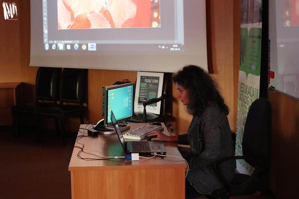 W audytorium, przy biurku zastawionym dwoma komputerami – stacjonarnym i laptopem, siedzi Adrianna Pohl-Gasidło. Kobieta o długich kręconych włosach, ubrana w ciemną sukienkę, z uwagą wpatruje się w ekran laptopa. Za prowadzącą widać fragment ekranu projektora, na którym wyświetla się trudne do zidentyfikowania zdjęcie.