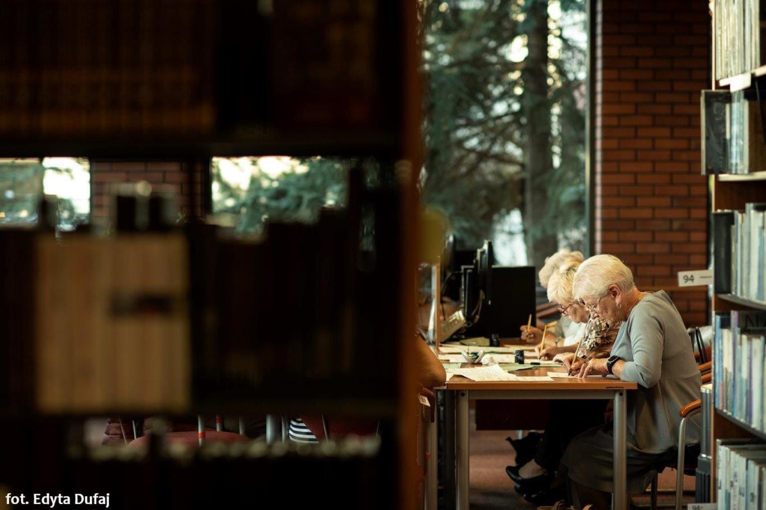Zdjęcie zrobione z oddali, zza regału z książkami w Czytelni Naukowej. Przy stole siedzą trzy siwowłose kobiety, a każda z nich pochyla się w skupieniu nad arkuszem, na którym starannie kaligrafuje ciągi znaków.