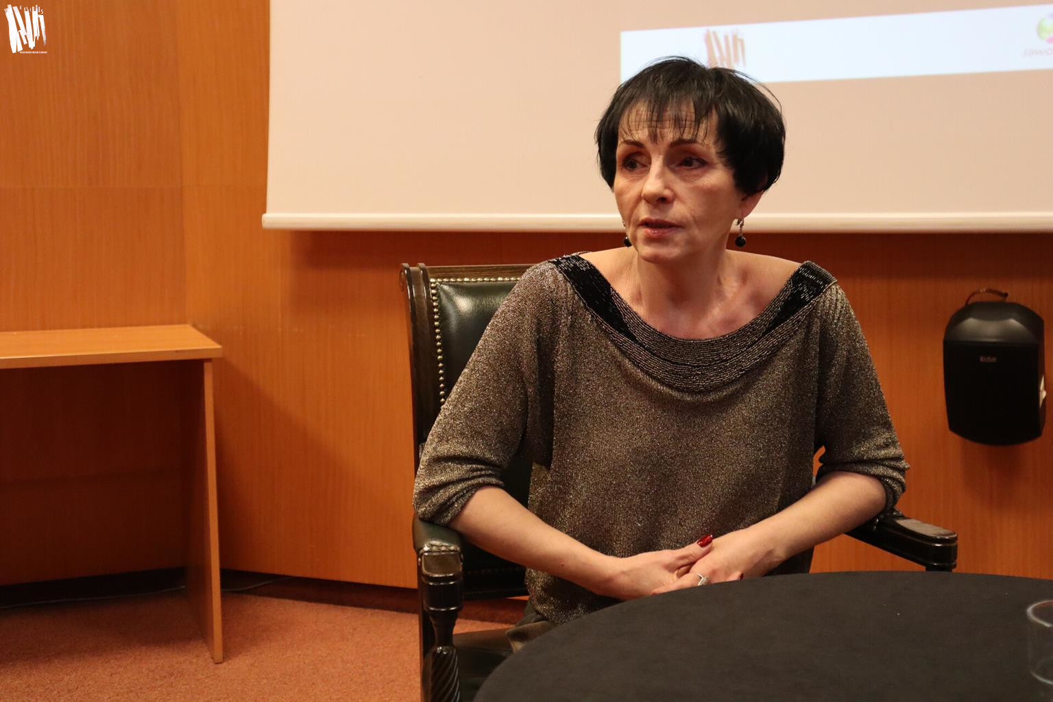 Audytorium Biblioteki Głównej, zbliżenie na Danutę Noszczyńską. Kobieta ma krótkie, ciemne włosy, ubrana jest w bluzkę dzianinową ze złotą nitką, okrągły dekolt wykończony ciemniejszą oblamówką.