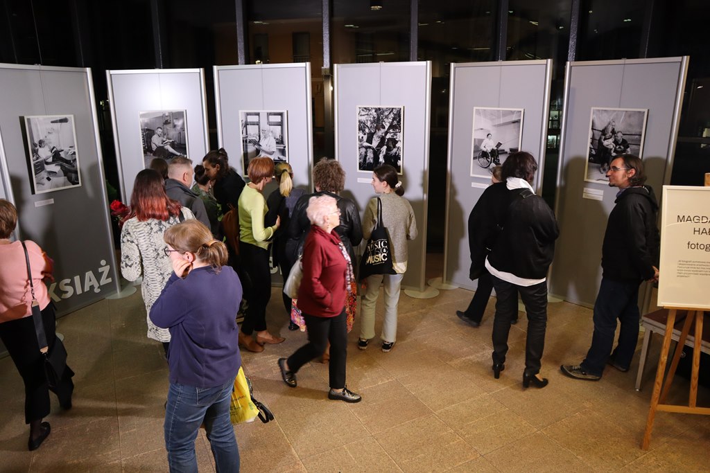 W przestrzeni Galerii ExLibris znajduje się piętnaście osób, które podziwiają czarno-białe fotografie wykonane podczas projektu.