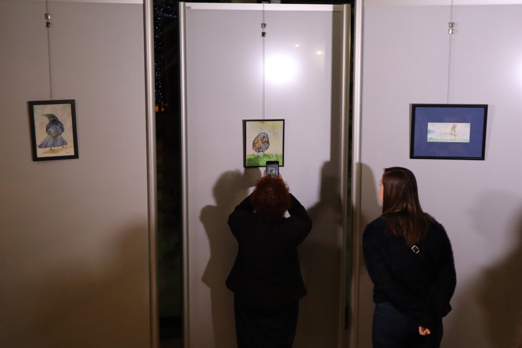 W Galerii ExLibris, odwrócone tyłem do kamery, stoją dwie osoby. Obie kobiety mają ciemne włosy i są ubrane na czarno, a ich uwagę pochłaniają niewielkie obrazy wyeksponowane w galerii. Kobieta stojąca po lewej stronie, za pomocą telefonu, robi zdjęcie jednego z obrazów.