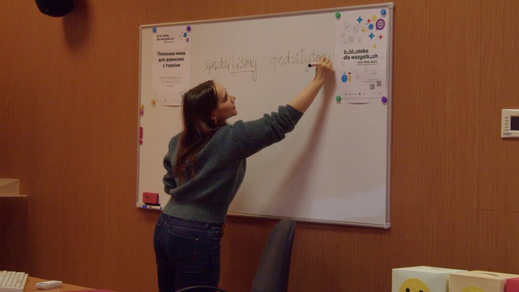 W audytorium młoda kobieta prowadzi zajęcia z języka polskiego. Nauczycielka stoi tyłem do kamery, pisząc na tablicy odmianę słowa „spędzać". Kobieta ma długie brązowe włosy, a ubrana jest w szary sweter i granatowe dżinsy.