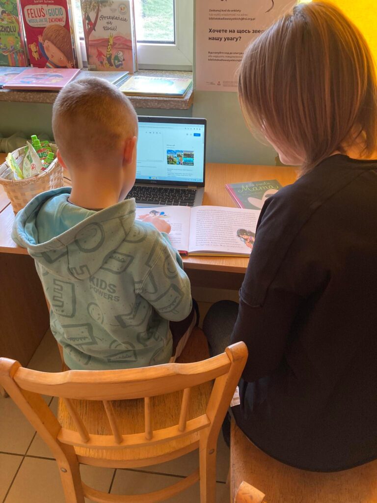 Tło: MBP Filia Podłęże 1. Przy stoliku pod oknem siedzą dwie osoby – kilkuletni chłopiec we wzorzystej ciemnozielonej bluzie oraz młoda kobieta ubrana na czarno. Czytelnicy siedzą tyłem do obiektywu, skupieni na czytaniu leżącej między nimi książki oraz informacjach wyświetlanych na niewielkim laptopie, który znajduje się bliżej chłopca. Częściowo widoczny parapet okna jest zastawiony książkami o różnokolorowych okładkach, a na ścianie nieopodal wisi plakat promujący projekt „Biblioteka dla wszystkich" w języku ukraińskim.