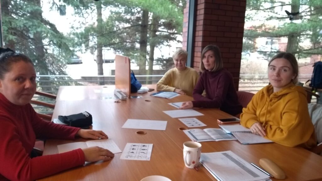 Czytelnia Naukowa. Przy długim stole, zasłanym materiałami do nauki języka polskiego, siedzą cztery kobiety. Uczestniczki zajęć z uśmiechem spoglądają w stronę kamery, a każda ma przed sobą tabelę z przydatnymi słówkami.
