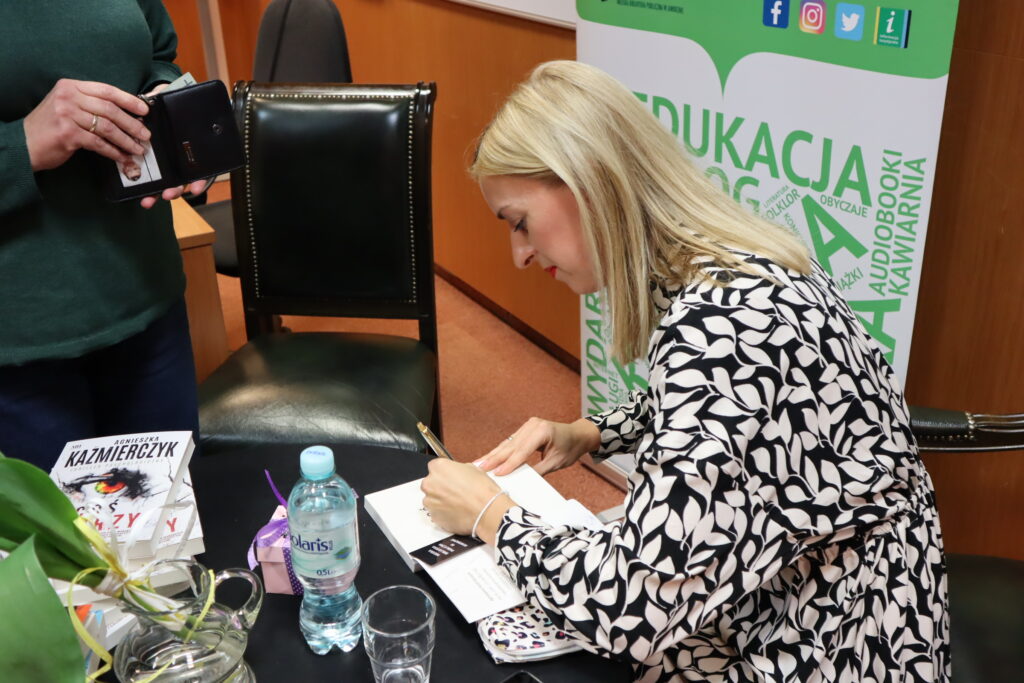 Autorka Agnieszka Kaźmierczyk, w czarno-białej sukience, składająca autograf na książce. Na czarnym stoliku sterta książek, butelka z wodą, szklanka, wazon z kwiatami. W tle krzesło, przy nim kobieta w zielonym swetrze i dżinsach z portfelem w ręku.