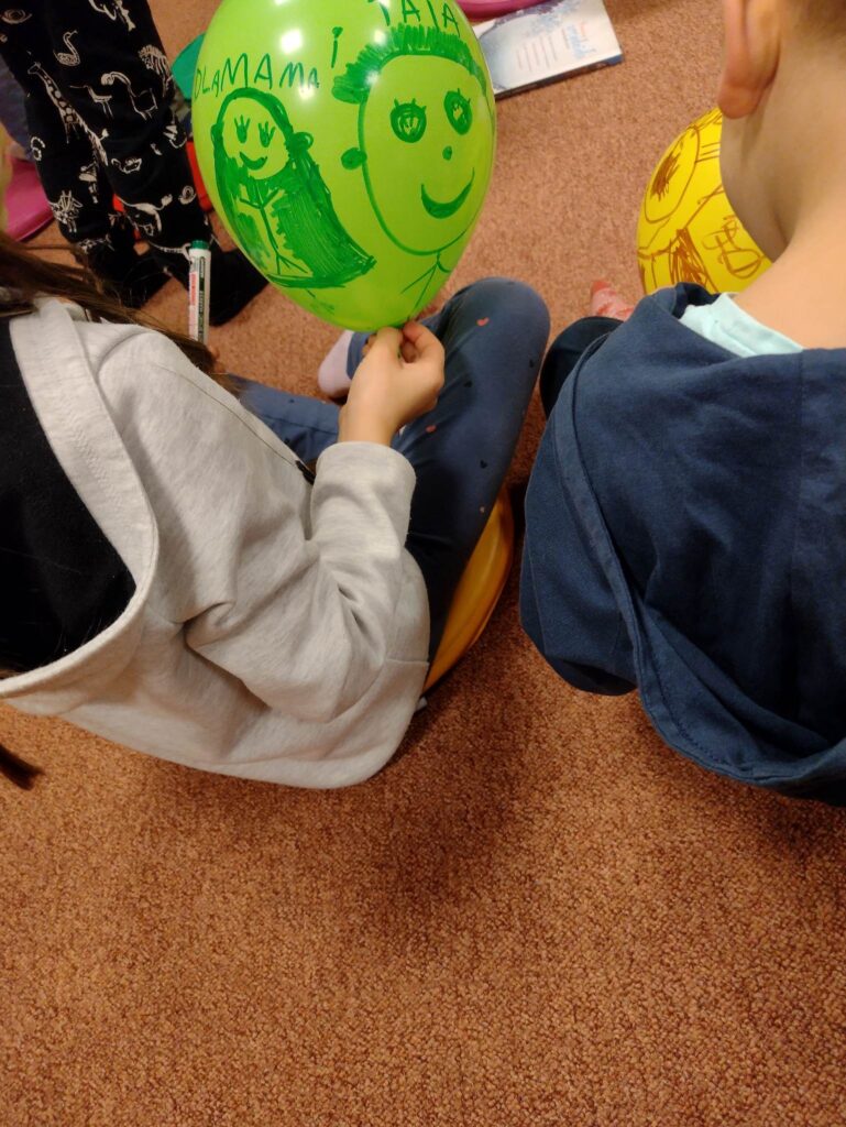 Sala ODM. Dzieci siedzą na kolorowych okrągłych poduszkach i rysują na balonach. Zielony balon, należący do uczestniczki siedzącej najbliżej kamery, został ozdobiony wizerunkiem jej mamy oraz twarzą jakiegoś chłopca – być może taty, brata, przyjaciela.