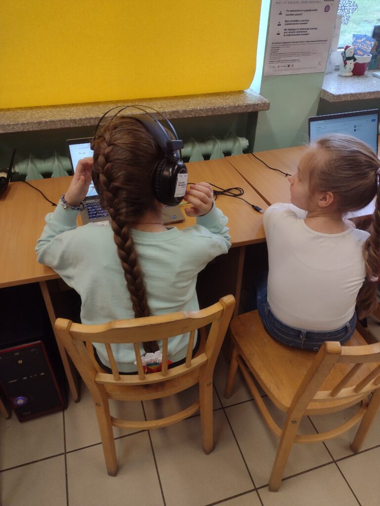 Przy stoliku, na terenie Filii Podłęże 1, siedzą dwie dziewczynki. Pierwsza, z bardzo długim brązowym warkoczem, ma na uszach słuchawki i w skupieniu wpatruje się w ekran stojącego przed nią laptopa. Siedząca obok młoda czytelniczka, o jasnych włosach spiętych w kucyk, nie ma na uszach słuchawek, ale i tak z zaciekawieniem zerka na ekran laptopa koleżanki. Pierwsza dziewczynka ma na sobie miętową bluzę i czarne spodnie, natomiast druga ubrana jest w białą bluzkę i granatowe dżinsy.