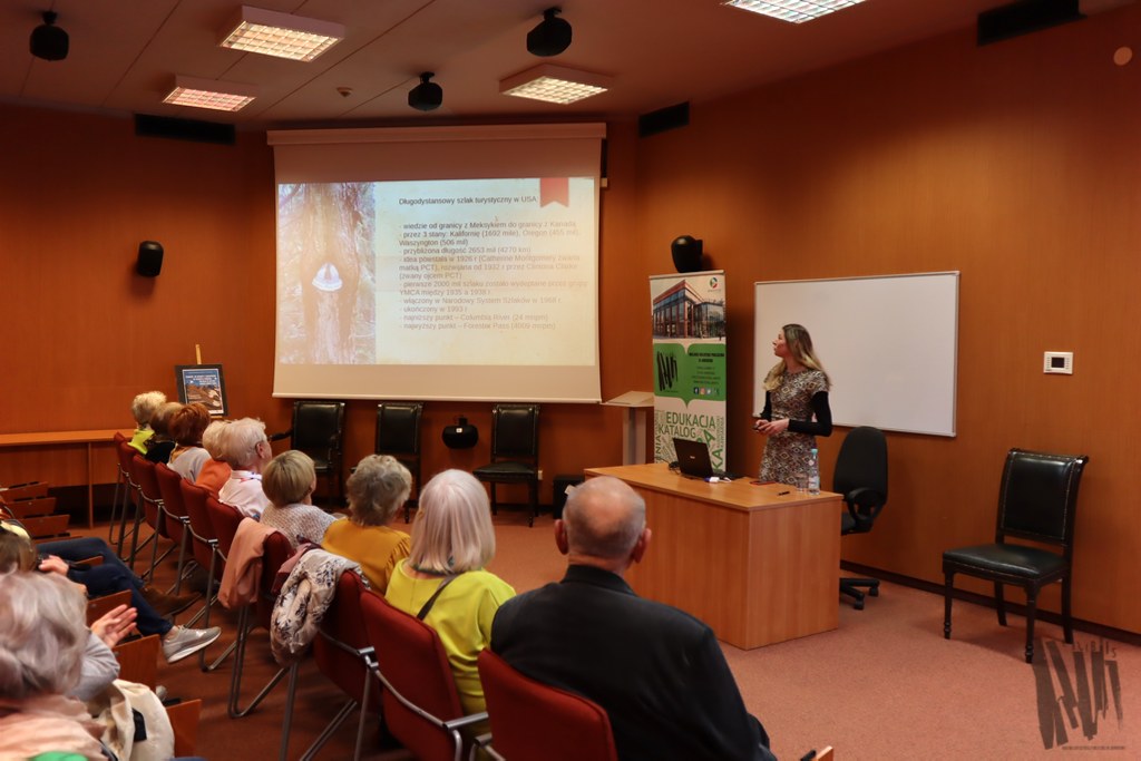 W audytorium, nieopodal ekranu projektora, na którym wyświetla się slajd pt. „Długodystansowy szlak turystyczny w USA”, stoi Kalina Namida. Kobieta ma długie blond włosy, a ubrana jest we wzorzystą czarno-białą sukienkę. Prowadząca patrzy na ekran z prezentacją i przemawia do publiczności zajmującej dwa rzędy krzeseł.