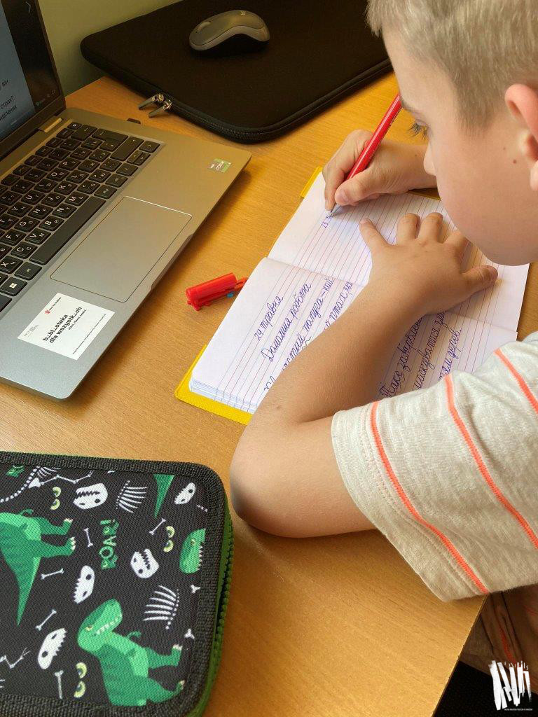 Przy biurku siedzi chłopiec w wieku szkolnym. Przed sobą ma włączonego laptopa, zapisuje coś w zeszycie w linie, zapis jest w języku ukraińskim. Po lewej stronie leży piórnik.