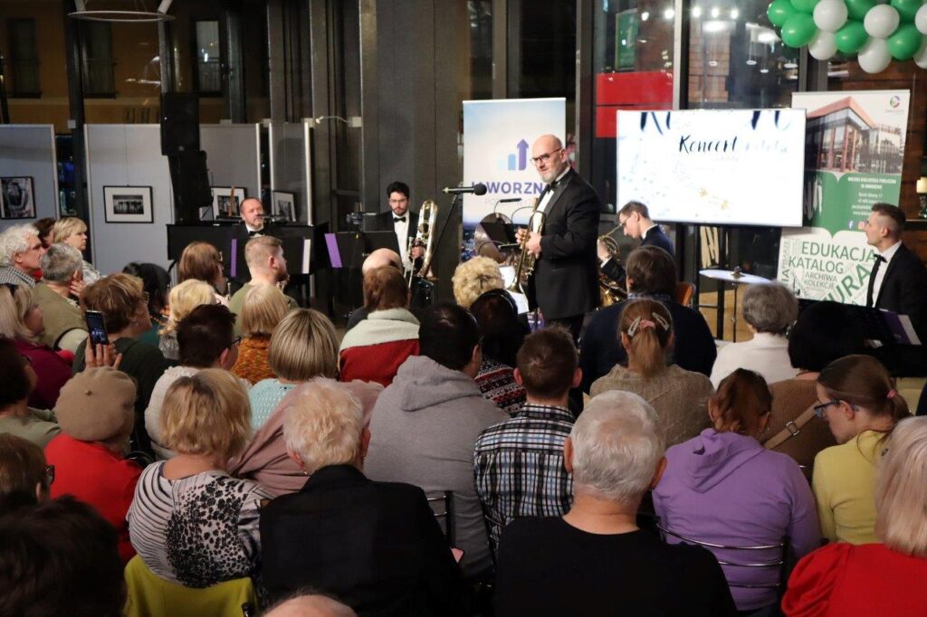 Na tle przeszklonych ścian Biblioteki Głównej stoi sześciu, elegancko ubranych, muzyków. Pięciu mężczyzn jest ustawionych nieco z tyłu, mając przed sobą podstawki na nuty. Jeden z muzyków, starszy mężczyzna, którego twarz zdobią okulary i siwa broda, stoi o krok z przodu i przemawia do tłumnie zgromadzonej publiczności.