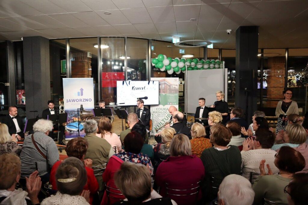 Tłumnie zgromadzona publiczność oklaskuje muzyków z zespołu eMBand. Obok muzyków, przy głównym mikrofonie, stoi bibliotekarka o krótkich jasnych włosach, ubrana w czerń. Nieco dalej stoi uśmiechnięta Dyrektor MBP, o ciemnych włosach spiętych w kok, ubrana w czarno-białą wzorzystą sukienkę. Za plecami występujących widać rollup Biblioteki, akcji Jaworzno poziom wyżej oraz ekran wyświetlający plakat reklamujący koncert.