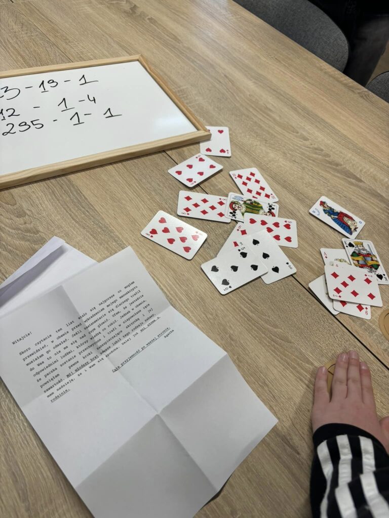 Zbliżenie na stolik, na którym znajdują się: wydruk zawierający zadanie dla uczestników, stos kart do gry oraz ramka z odręcznie zapisanymi działaniami matematycznymi. 