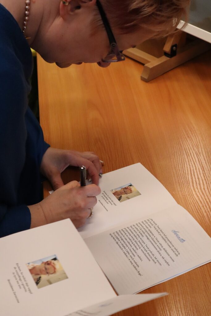 Na kolorowym zdjęciu widzimy pochyloną kobietę w średnim wieku o jasnych włosach w niebieskiej bluzce z koralami na szyi i okularach, która trzyma w prawej dłoni długopis, którym podpisuje tomik poezji. Na biurku leży drugi rozłożony tomik, przygotowany do podpisu.