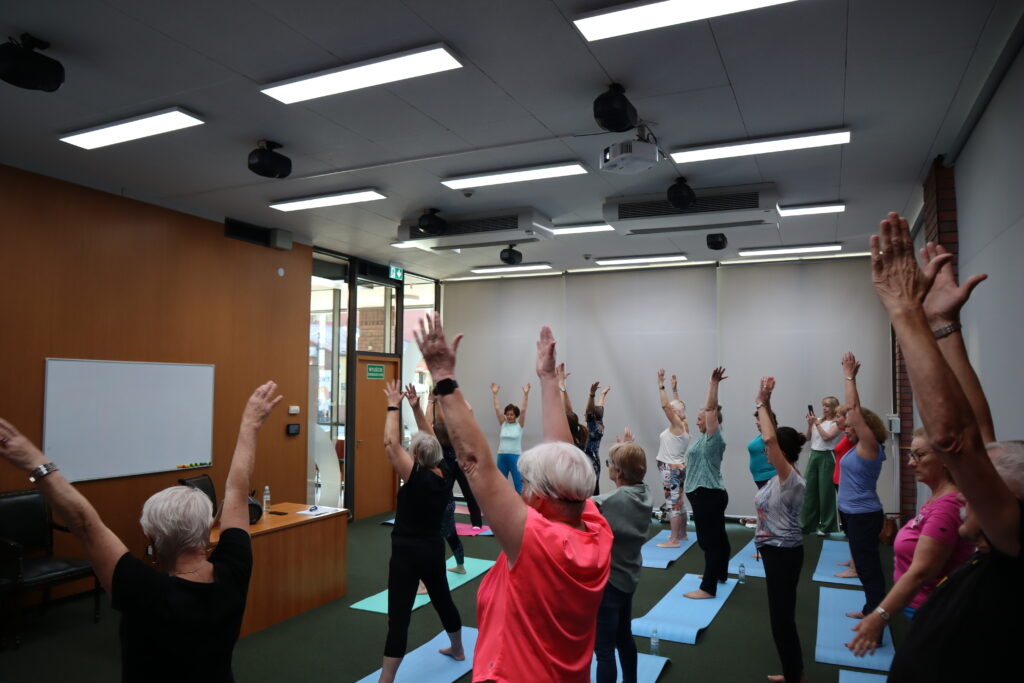 Seniorzy, ubrani w kolorowe stroje sportowe, stoją na matach w przestrzeni audytorium. Uczestnicy wykonują ćwiczenie polegające na rozciąganiu ciała i wyciąganiu rąk w stronę sufitu.