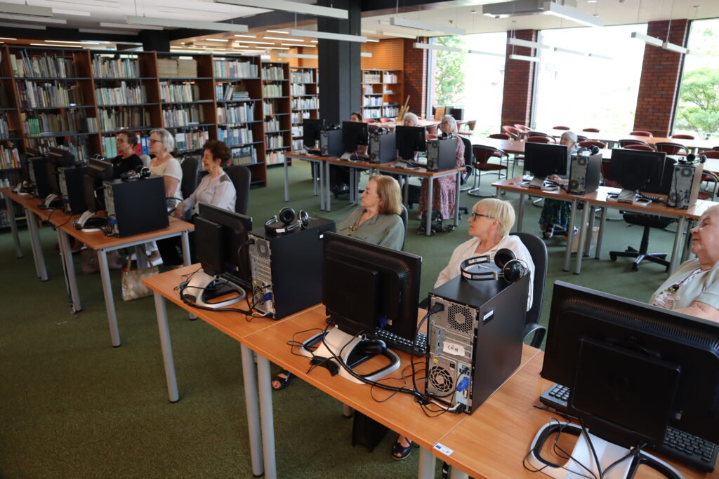 W Czytelni Komputerowej, przy uruchomionych urządzeniach, siedzi dziesięć seniorek. Kobiety z zainteresowaniem śledzą wykład i poczynania niewidocznego Patryka Mataniaka, edukatora prowadzącego warsztaty.