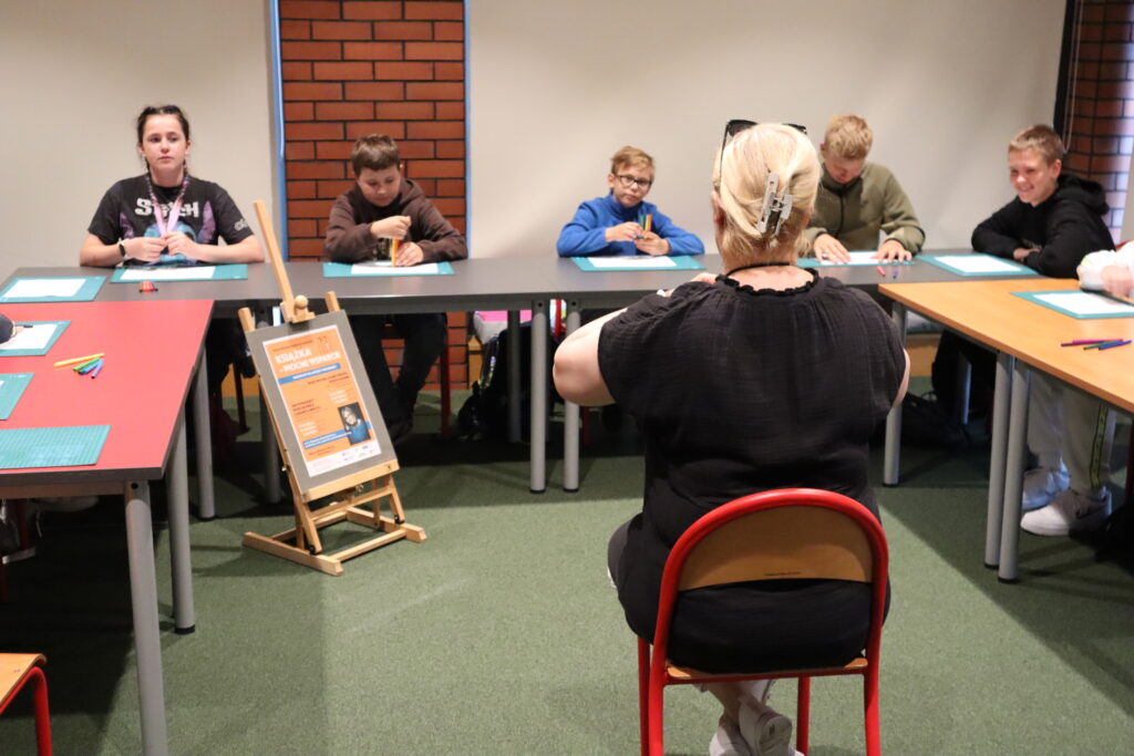 Przed dziećmi uczestniczącymi w warsztatach siedzi Magdalena Dziedziak-Wawro. Jasnowłosa kobieta, ubrana w czarną tunikę i białe spodnie, siedzi tyłem do obiektywu i czyta książkę pt. „Alfabet szczęścia z coachingiem w tle i kotem na kolanach”. 