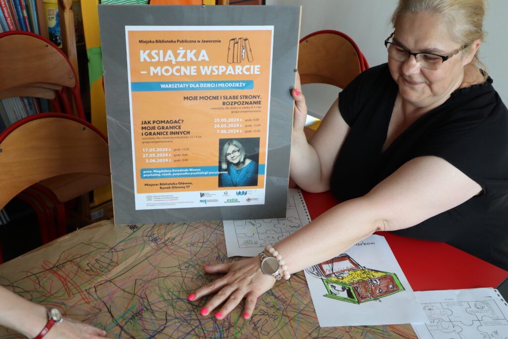 Przy plakacie promującym warsztaty siedzi Magdalena Dziedziak-Wawro. Jasnowłosa psycholożka, której twarz zdobią okulary, ubrana jest w czarną bluzkę i czerwone spodnie. Uśmiechnięta kobieta trzyma dłoń na pracach dzieci, czyli skrzyniach skarbów i pracy zbiorowej wykonanej na szarym papierze. 