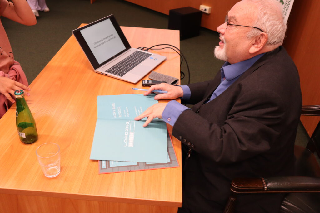 Zbliżenie na biurko, przy którym siedzi prof. Ryszard Tadeusiewicz, podpisujący książkę dla kilkuletniej dziewczynki. Na biurku widać również laptop, na ekranie którego wyświetla się slajd prezentacji pt. „Oby sztuczna inteligencja była w naszych rękach – a nie my w jej!".